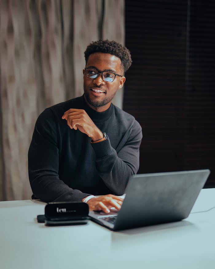 about-us-02 Cheerful man with glasses working on laptop at a table indoors, emanating a professional and modern workspace vibe.